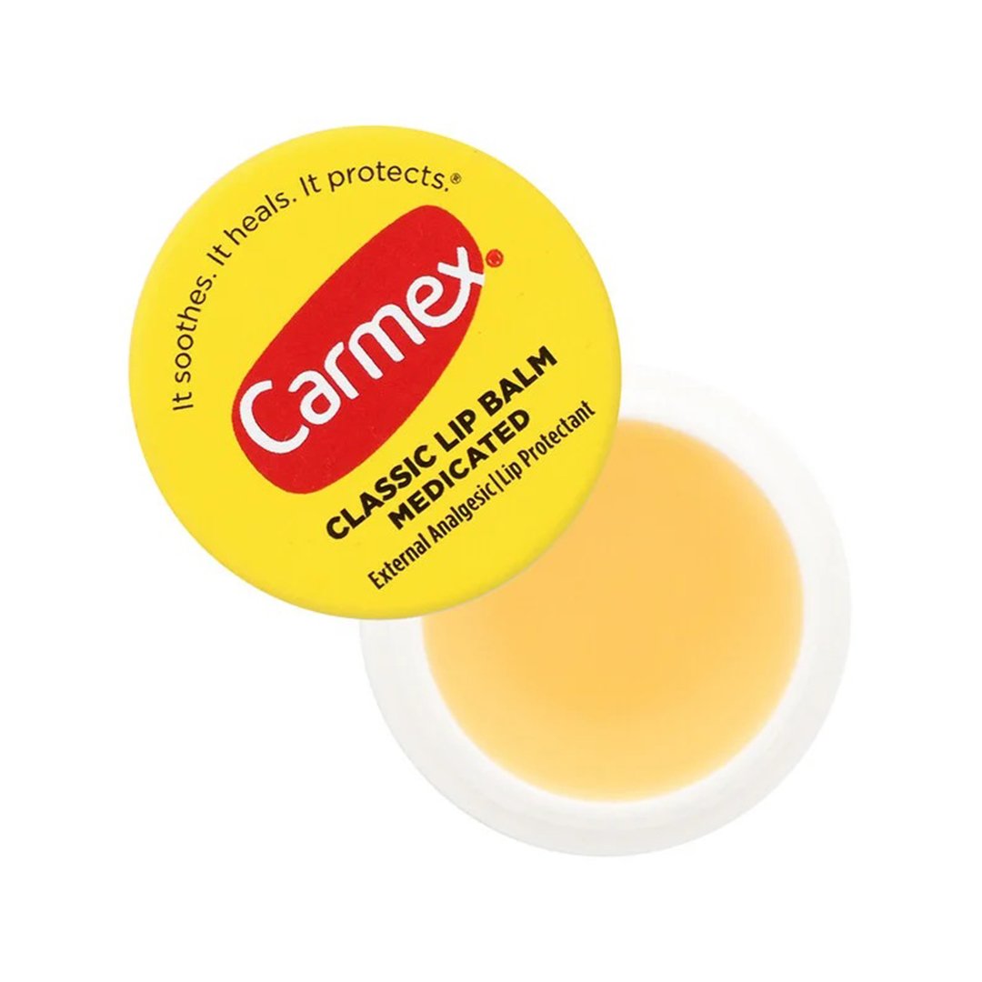 A yellow tin of Carmex Classic Medicated Lip Balm featuring the brand's red logo. Text on the lid reads: "It soothes. It heals. It protects.